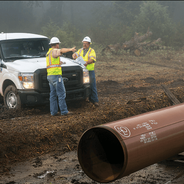 Image of an Atmos Energy truck with a door open, with 2 workers in front of the truck pointing to a work area with a large pipe